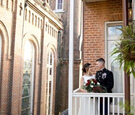 Bride and groom on a balcony, holding a bouquet, gaze at each other with a tall brick church in the background.