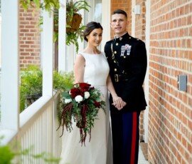 Bride in white dress with bouquet stands beside groom in military uniform on brick porch, classic Gulf South wedding scene.