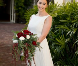 Bride in white dress on a brick path, holding red and white bouquet, smiling at her New Orleans wedding.