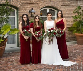 Bride in white and three bridesmaids in red pose with bouquets by brick walls and potted plants at a Louisiana wedding.