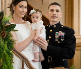 Bride, groom in military uniform, and baby smile on a greenery-adorned staircase at their elegant Gulf South wedding.