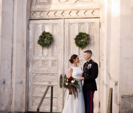 Bride in white gown and groom in uniform hold hands on church steps with wreaths, during a classic New Orleans wedding.