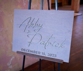 Wooden wedding sign on easel with “Abby + Patrick” and “December 15, 2023” in gold script, rustic indoor setting.