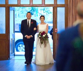 Bride in white dress and bouquet enters room arm-in-arm with suited man, blurred guests in foreground at wedding ceremony.
