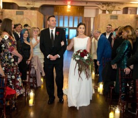 Bride in white dress walks down aisle with man in black suit at warmly lit Gulf South wedding as smiling guests look on.