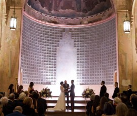 Elegant New Orleans wedding in a grand hall with tall arches, mural ceiling, and candlelit aisles filled with guests.