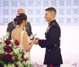 Bride and groom in military uniform exchange rings at Gulf South wedding, officiant reading, red and white flowers in front.