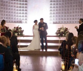 Bride and groom holding hands and smiling at a beautifully decorated indoor Gulf South wedding ceremony with guests, flowers, and candles.