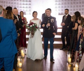 Bride in white dress and groom in military uniform walk down aisle, smiling, at elegant Louisiana wedding with candles.