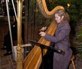 Woman in a purple coat plays ornate golden harp on a nighttime patio, wedding inspiration with music sheets and lush plants.