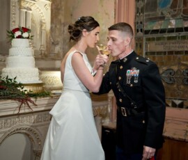 Bride in white dress and groom in military uniform toast beside a white cake with red and white flowers at their Gulf South wedding.