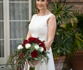 Bride in white dress with red and white bouquet, smiling outdoors by plants and brick wall—Louisiana wedding inspiration.