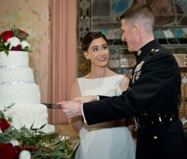 Bride and groom, in military dress and white gown, cut a white cake surrounded by red and white flowers at Gulf South wedding.