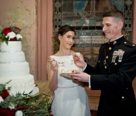 Bride and groom, with the groom in military uniform, share cake at their Louisiana wedding beside a white cake with red flowers.