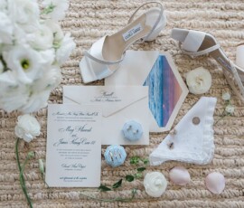 Elegant wedding flat lay: white heels, invites, bouquet, beach photo, ring box, jewelry, and seashells on woven mat.