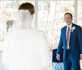 Groom in blue suit smiles at bride in white dress and veil, her back to camera indoors—Gulf South wedding inspiration.