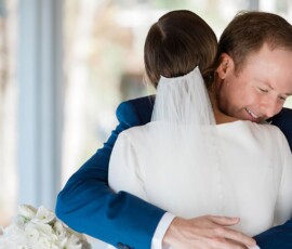 Bride and groom embrace indoors at their elegant New Orleans wedding; she holds white flowers, he smiles with eyes closed.