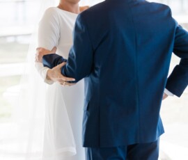 Bride in white dress and man in blue suit smile and hold hands at indoor wedding, sunlight shining through large windows.