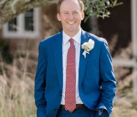 Man in blue suit with red patterned tie and boutonniere smiles outdoors, greenery and building behind; stylish New Orleans wedding inspiration.