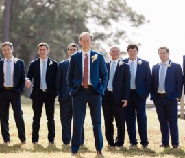 Smiling groom with groomsmen in suits and white boutonnieres at a sunny outdoor Louisiana wedding with trees behind them.