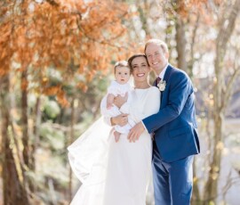 Smiling couple in formalwear holds a baby among autumn trees; she wears white, he blue, with sunlit leaves in the background.