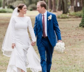 Bride in white dress and veil walks hand-in-hand with groom in blue suit outdoors, smiling after wedding near rustic New Orleans building.