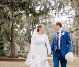 Bride in long-sleeved white gown and groom in blue suit smile, holding hands outdoors with moss-draped trees in New Orleans.
