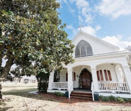 Small white church with arched windows and front porch under a large tree on a bright, partly cloudy Gulf South day.