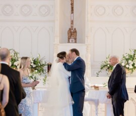 Bride and groom kiss at the church altar, surrounded by guests, flowers, and a large cross in the background.