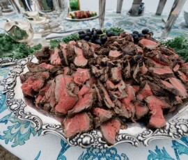 Medium-rare roast beef on a silver platter with greens and grapes at a wedding buffet table.