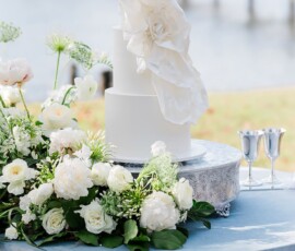 Elegant three-tier white wedding cake with floral decor on silver stand, blue tablecloth, and silver goblets by the water.