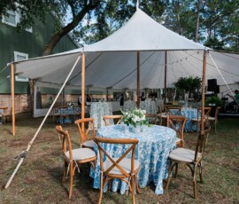 Outdoor wedding tent with blue-patterned tables, wooden chairs, floral centerpieces, and a green building in the background.