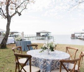 Round table with floral cloth and flowers, wooden chairs on grass by a lake with docks, trees, and partly cloudy sky.