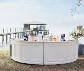 Curved white outdoor bar with liquor bottles by a lake, perfect Gulf South wedding inspiration with docks and blue sky backdrop.