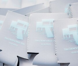 Light gray drink koozies with a white house and waves graphic, marked THE CROWS NEST TOO 12.9.2023 in light blue.