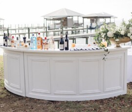 Outdoor curved white bar by the water with bottles, glasses, floral arrangement; docks and pavilions in background.