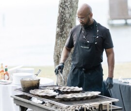 Man in apron and gloves grills oysters by a waterfront, with sauces and utensils—ideal Louisiana wedding inspiration.