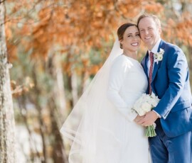 Bride in white dress and groom in blue suit embrace outdoors amid autumn trees, holding white bouquet at romantic Gulf South wedding.