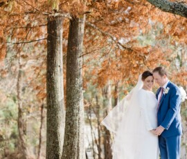 Bride in white and groom in blue suit smile together under autumn trees with sunlit orange leaves.
