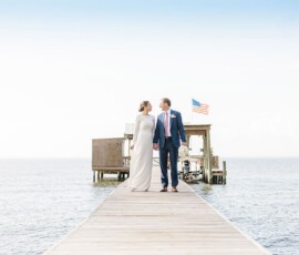 Bride and groom smile, holding hands on a wooden dock over water, with building and American flag in the background.