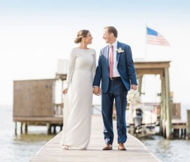 Bride and groom smiling and holding hands on a dock, with an American flag and building behind them at their New Orleans wedding.