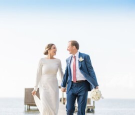 Bride in white dress and groom in blue suit hold hands by the water, smiling under a clear sky with docks behind.