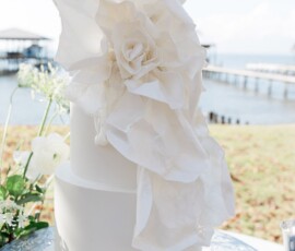 White two-tiered wedding cake with a large sugar flower, set outdoors by water and greenery—ideal for Louisiana weddings.