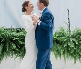 Bride in white dress and groom in blue suit dance and smile at their New Orleans wedding, surrounded by ferns and white curtains.