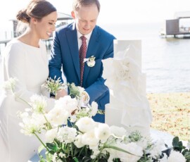 Smiling bride and groom cut a tall white wedding cake with flowers at an outdoor Gulf South waterfront reception.