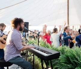 Musician plays keyboard and sings while another plays guitar; lively crowd dances under fern-decorated white tent at Louisiana wedding.