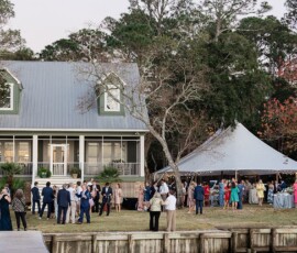 Guests in formal attire gather by a two-story house and white tent for an outdoor Gulf South wedding near a wooded dock.