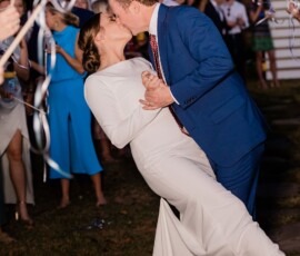Bride in white dress and groom in blue suit kiss as he dips her, with guests waving streamers at their New Orleans wedding.