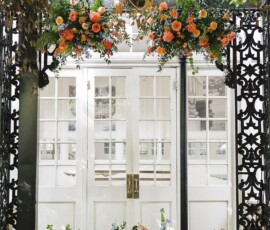 Elegant white double doors with glass panes, ornate black ironwork, lush florals, and a lantern—ideal Gulf South wedding backdrop.