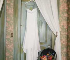 White wedding dress on a door with white curtains, colorful bouquet on black chair, vintage room with green trim and floral wallpaper.